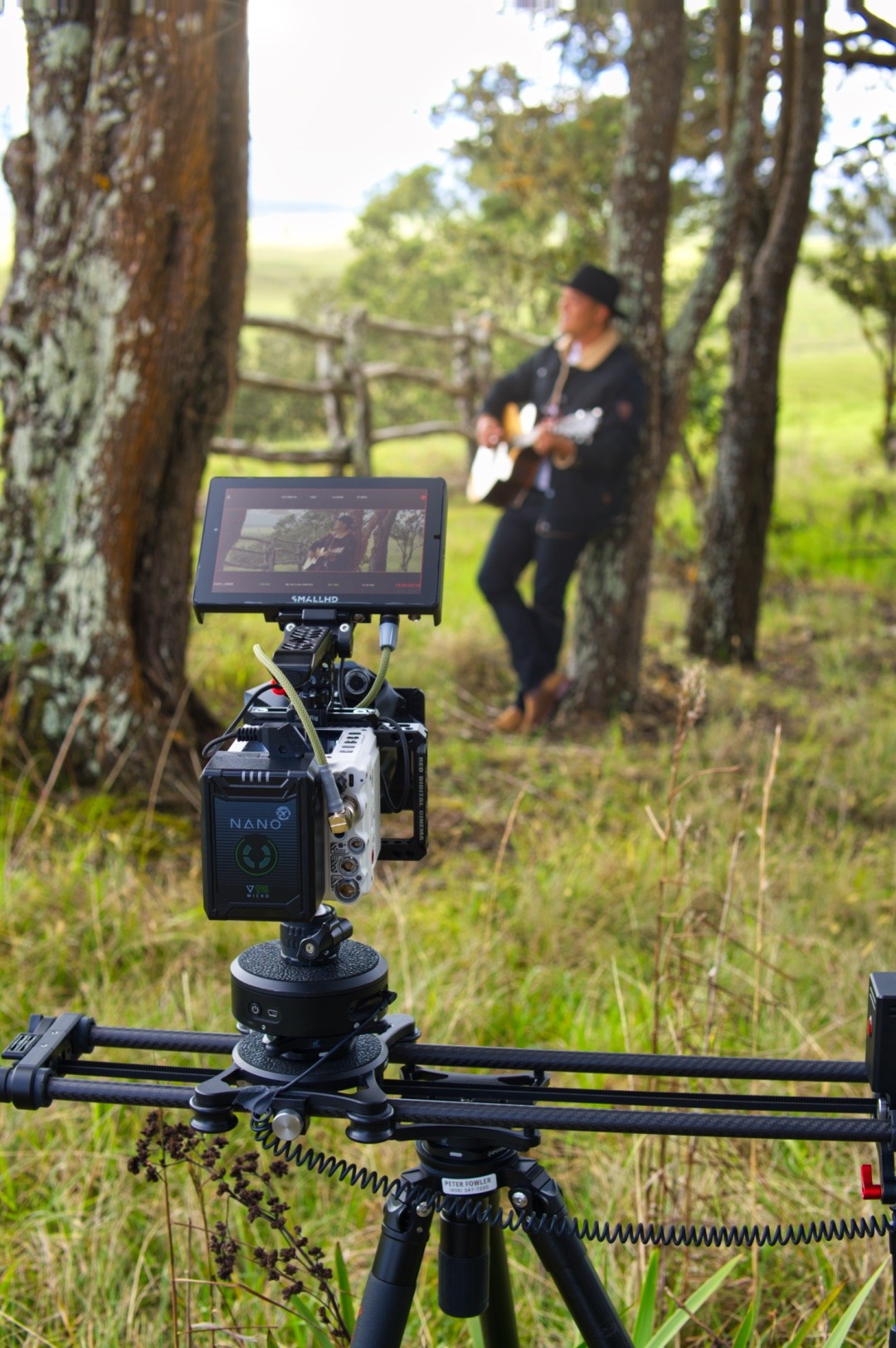 A cinema camera on a slider in tall grass, a guitarist standing among the trees behind it.